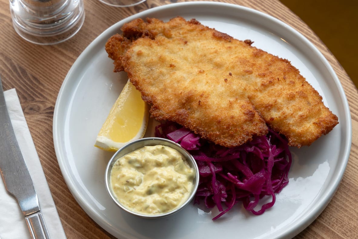 crumbed fish schnitzel on a plate with pickled red cabbage, a wedge of lemon and portion of tartare sauce.