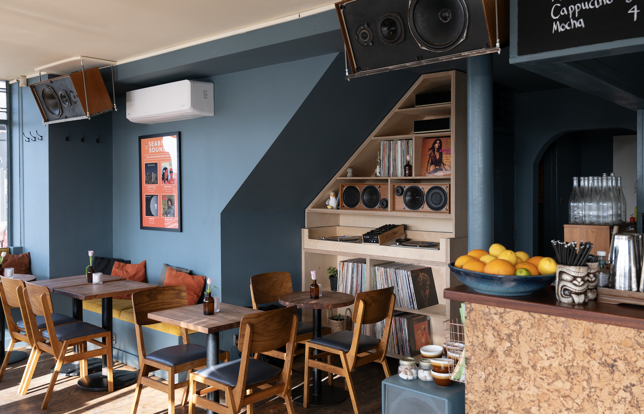 Cafe interior with wood seats and a pale teal wall with records, speakers and a bar in view.
