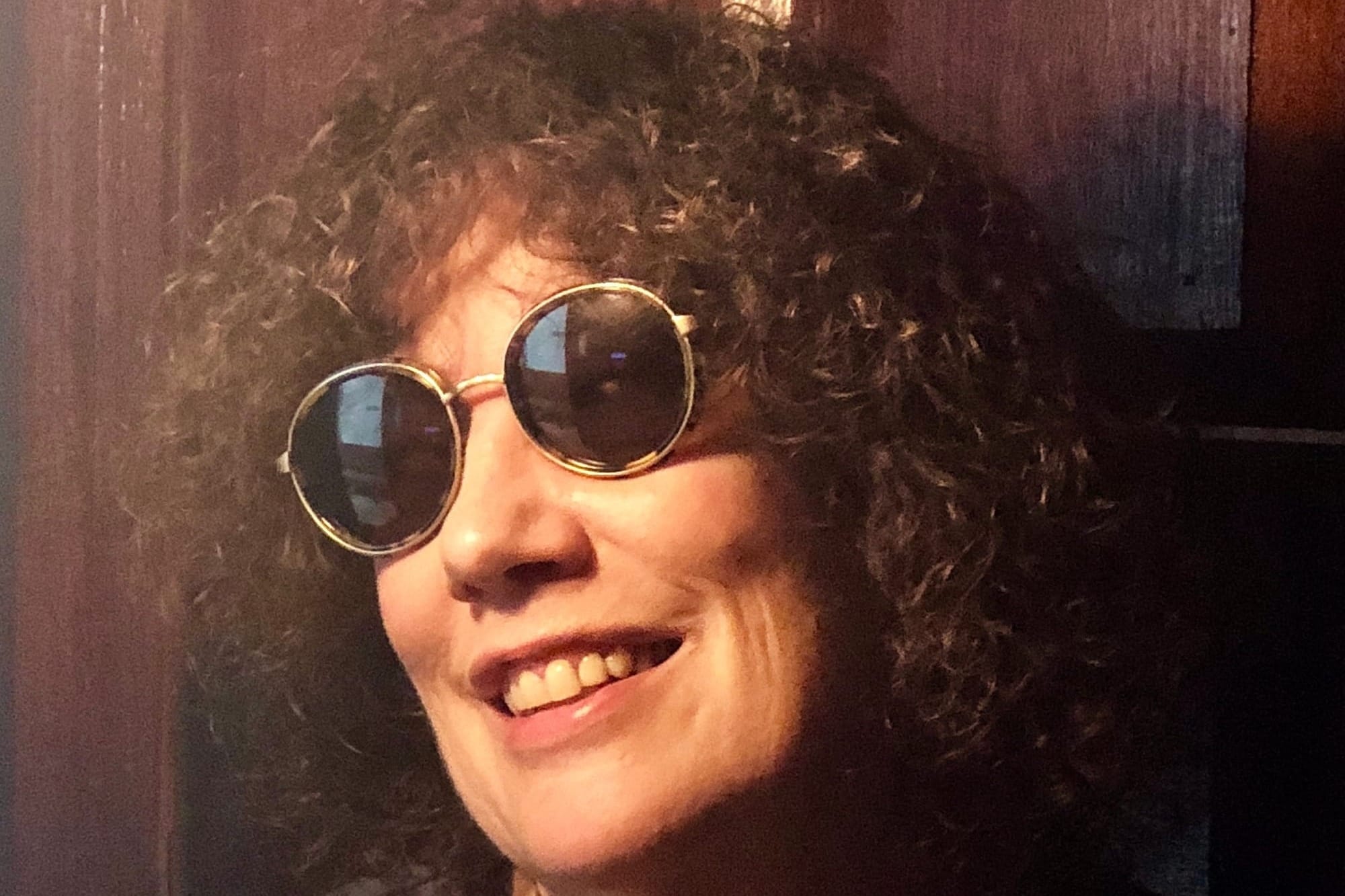 A woman with curly hair wearing round sunglasses, smiling.