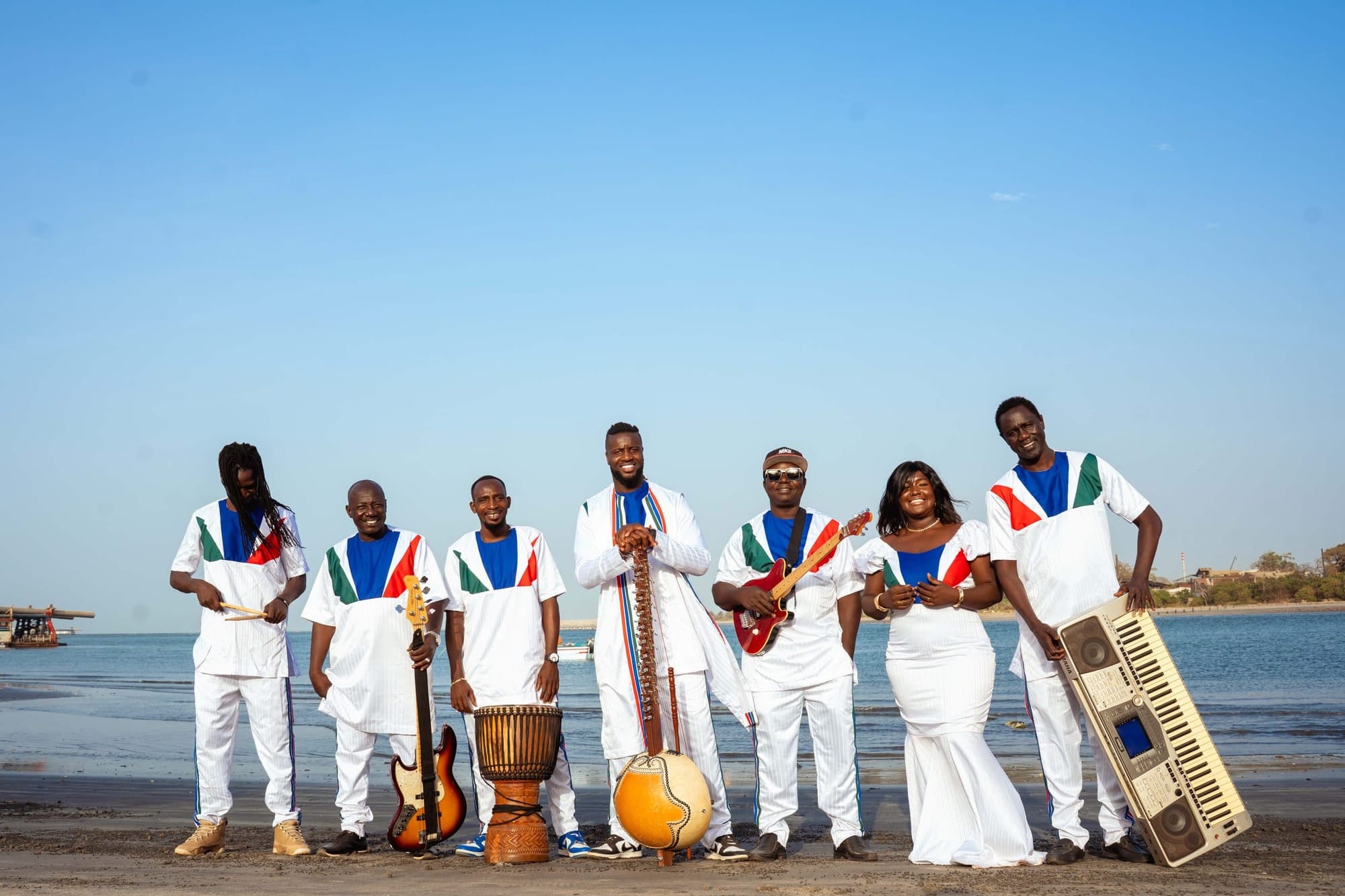 Seven band members posing in front of a body of water wearing white and holding traditional African instruments.