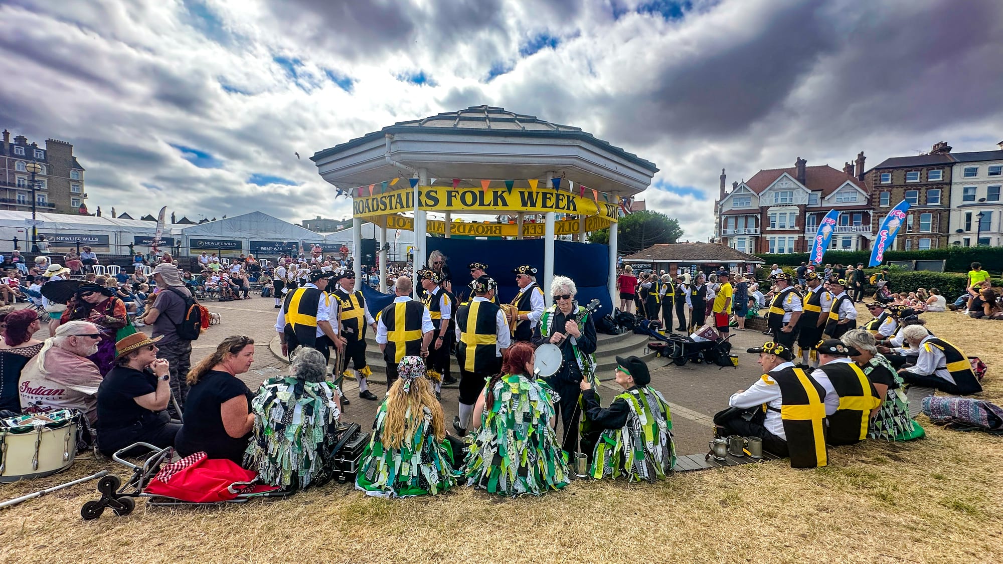 People dressed in black and yellow smocks and green tassled vests sit on the grass around a band stand