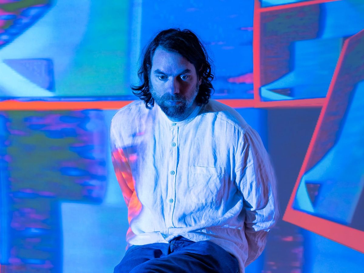 A man with dark hair and a beard wearing a white shirt sits as images are projected onto him and the wall behind him