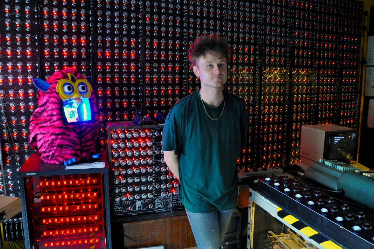 A man in a black t-shirt stands in front of a large back on dial and red LEDS. A large Furby is to the left of the frame.