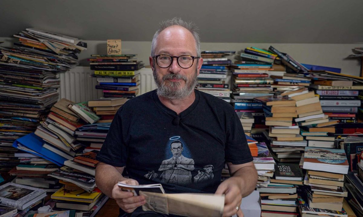 A man with grey hair and a bear sits in front of stack of books