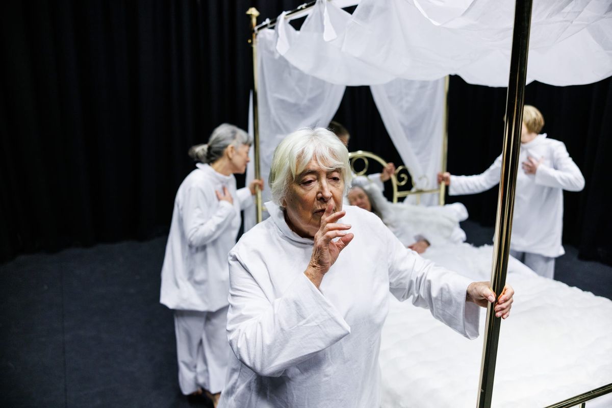 Several elderly women dressed in white stand around a gold four poster bed. The woman in the foreground holds her finger to her lips.