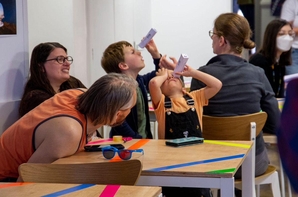 A group of people sit at tables. Two children look through kaleidoscopes.