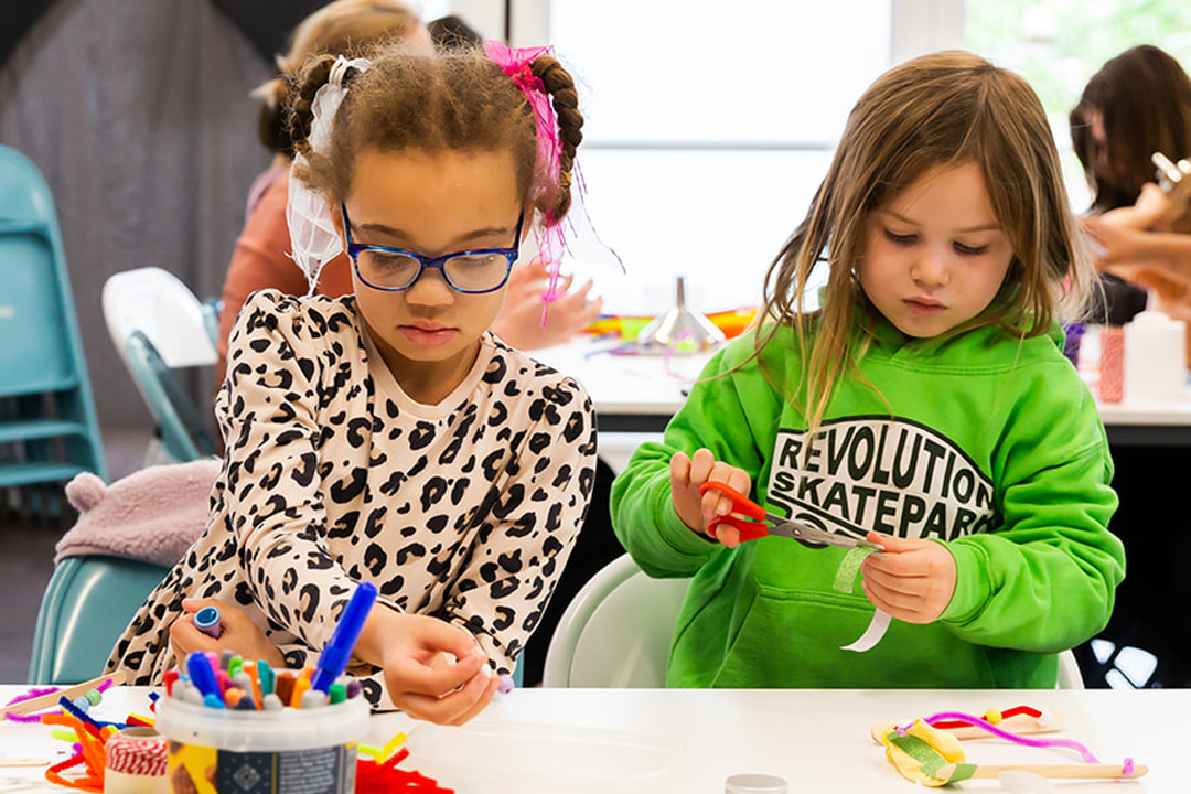 Two children site at a table doing crafts, one wears glasses and where's a leopard print top, the other wears a green hoodie reading 'Revolution skatepark'