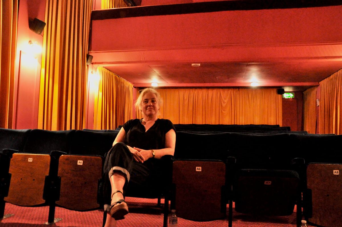 A woman with white hair sits in the front row of an otherwise empty cinema looking directly into the camera
