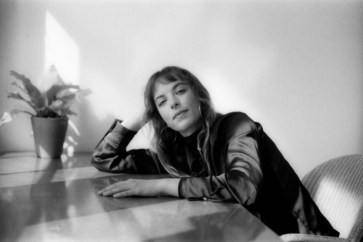 A black and white photo of a young woman with dark hair leaning with one elbow on a table and hand agains the back of her head. 