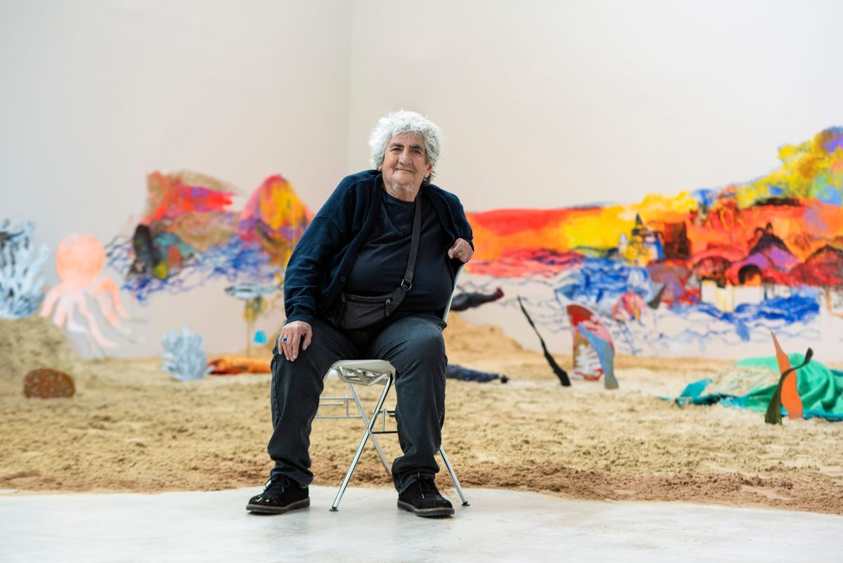 An elderly woman with grey hair sits on a stool, dressed in black with colourful art in the background