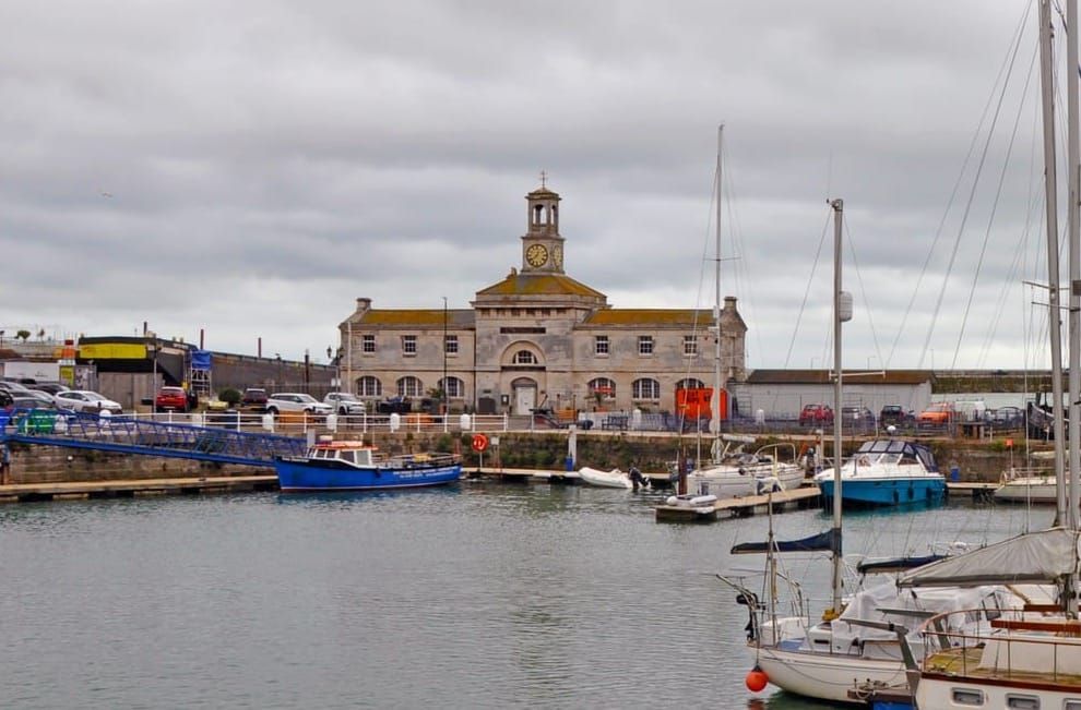 A building with a clock on top of it stands in front of a body of water in which boats can be seen.