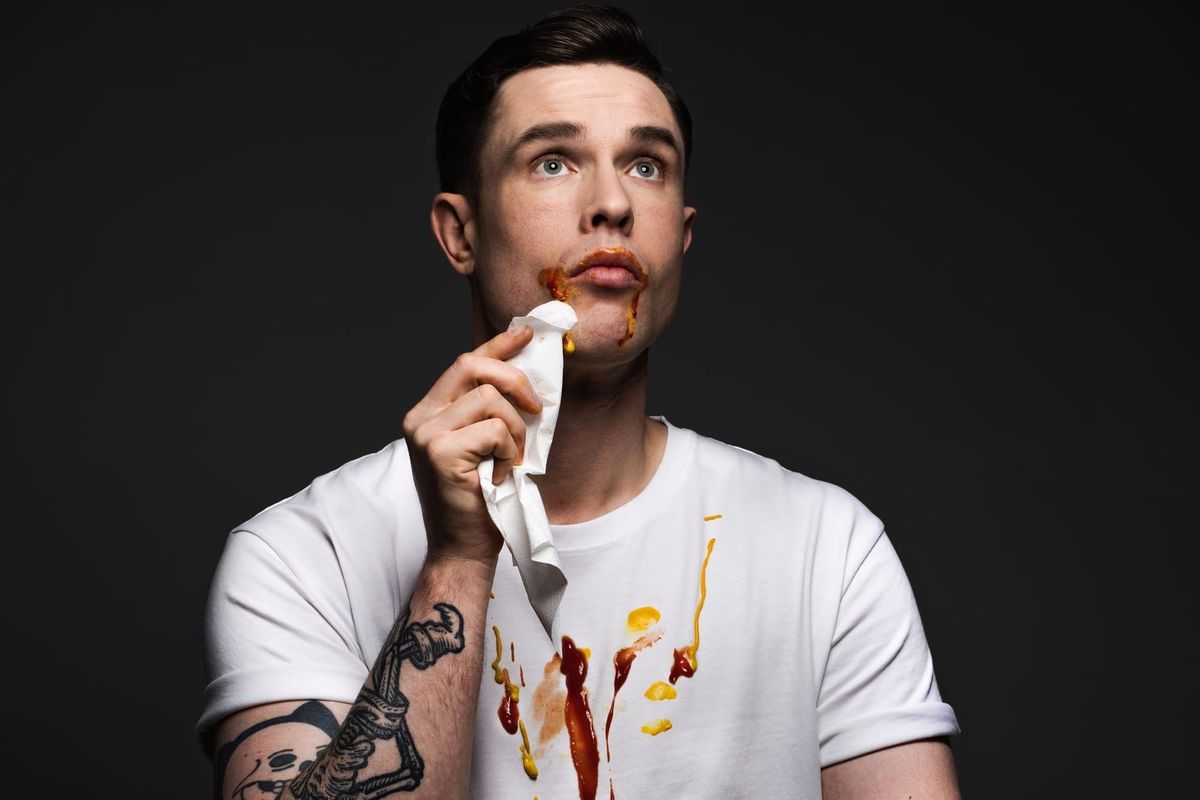 A man dabs at his face lightly with a napkin. His face and white t-shirt are splashed with mustard and ketchup.