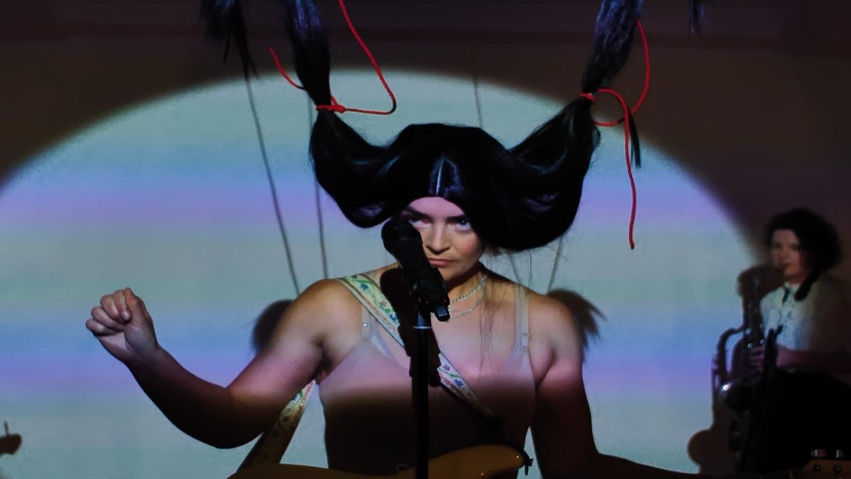 A woman stands behind a microphone wearing a bra. She wears a black wig with pigtail lifted up by red rope. Another woman plays saxophone in the background.