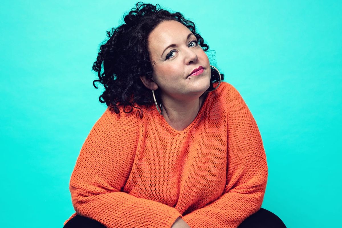 A woman with black hair, wearing hoop earrings, an orange knitted jumper and black trousers crouches with her head cocked to the side. 