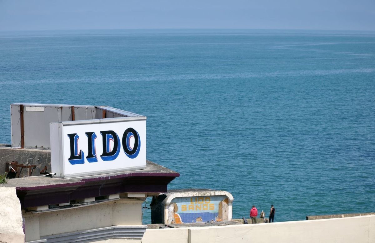 A view of the sea. In the foreground a sign reads 'LIDO'. In the distance another sign reads 'LIDO SANDS'. Two men stand by this second sign fishing. 