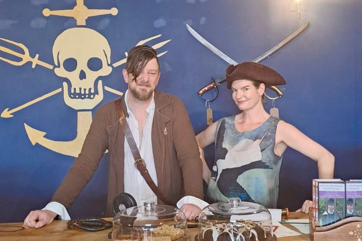 A man and a woman dressed as pirates stand in front of a skull and crossbones, and two crossed swords.