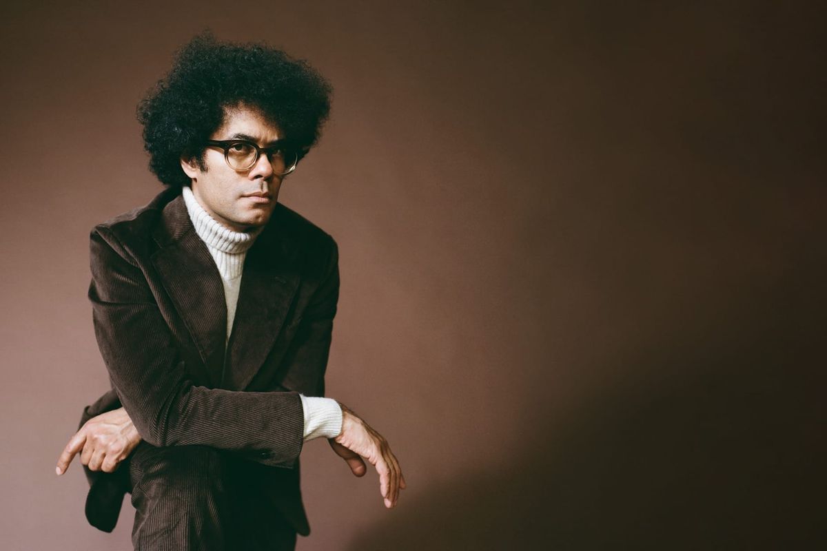 A man with black hair wearing a brown suit and white polo neck jumper against a brown background