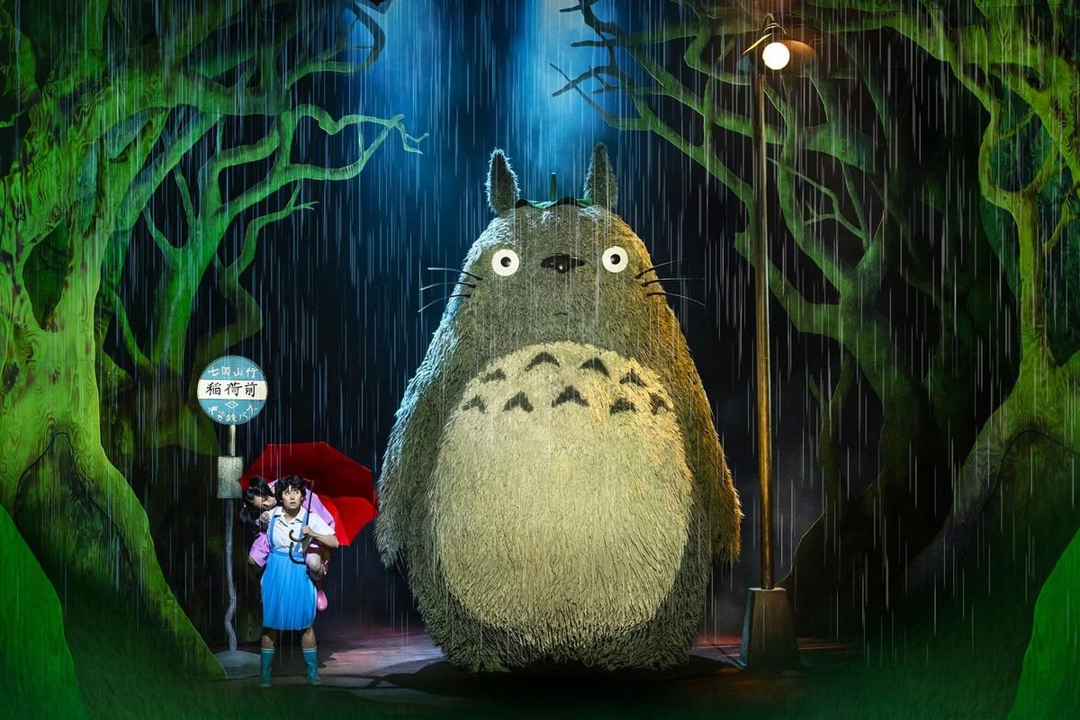 A large grey and white creature stands in the rain with a leaf on its head. Next to it are two girls, one on the others back, with a red umbrella. There is a bus stop.