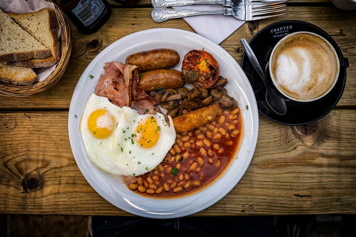 A British fry up on a white plate on a rustic wooden table with fried eggs, baked beans, sausages, bacon, mushrooms, and tomato. There is also a cup of coffee, a basket of bread and two forks