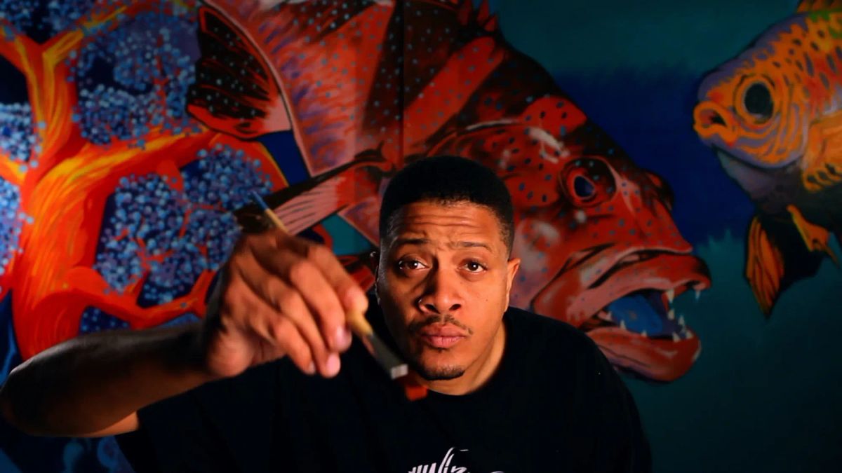 A man reaches up to the camera with a paintbrush. Behind him is a large painting featuring fish.