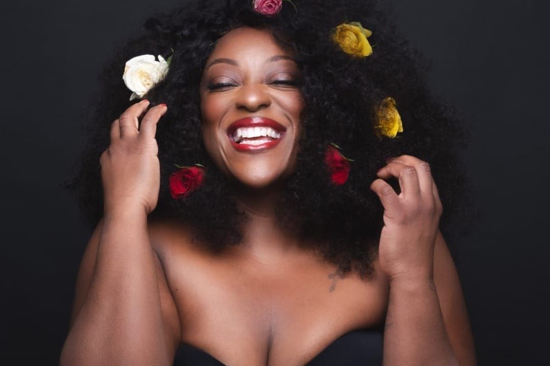 A woman wearing an off the shoulder dress laughs. She has on red lipstick. She has white, yellow and red roses in her hair. 