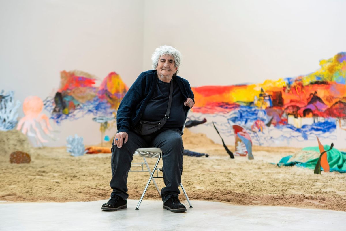 A woman in her 80s sits on a chair in front of a large art installation in a white walled room