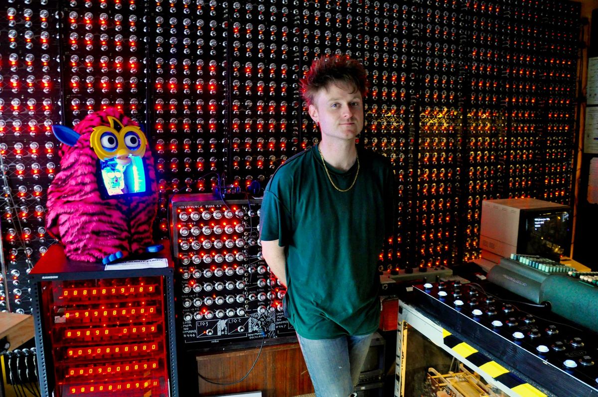 A man stands in front of a bank of red lights with a large Furby next to him