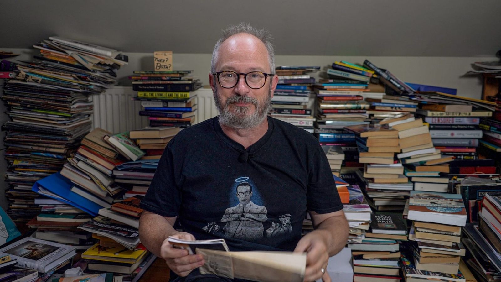 A beared man seated in front of piles of books