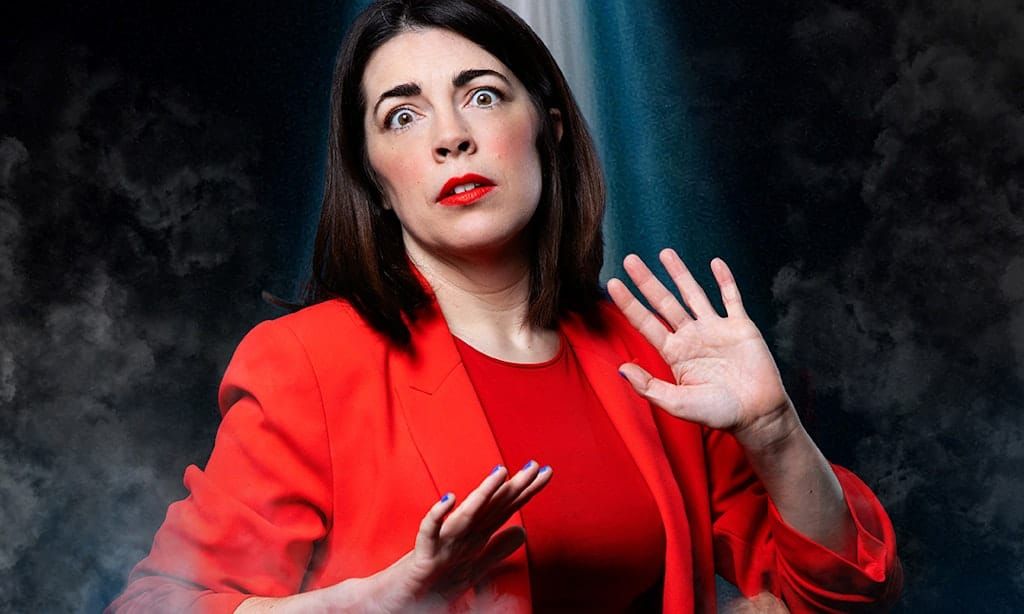 A women with brown hair wearing a red suit holds her hands up in shock