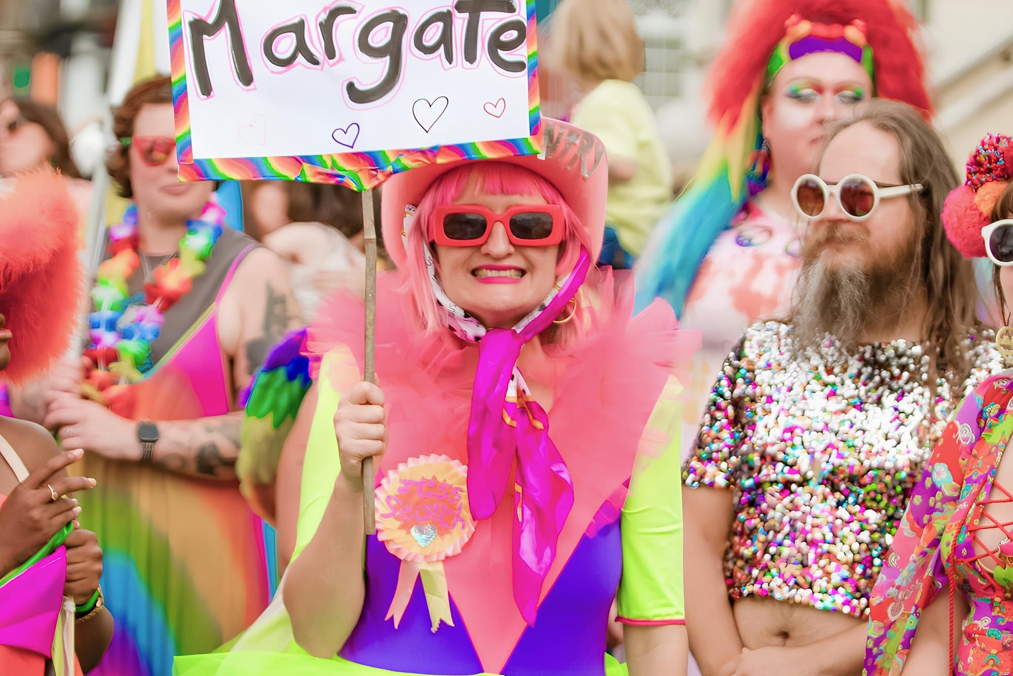 A group of people in colourful outfits. A smiling woman in the foreground holds a sign reading 'Margate'