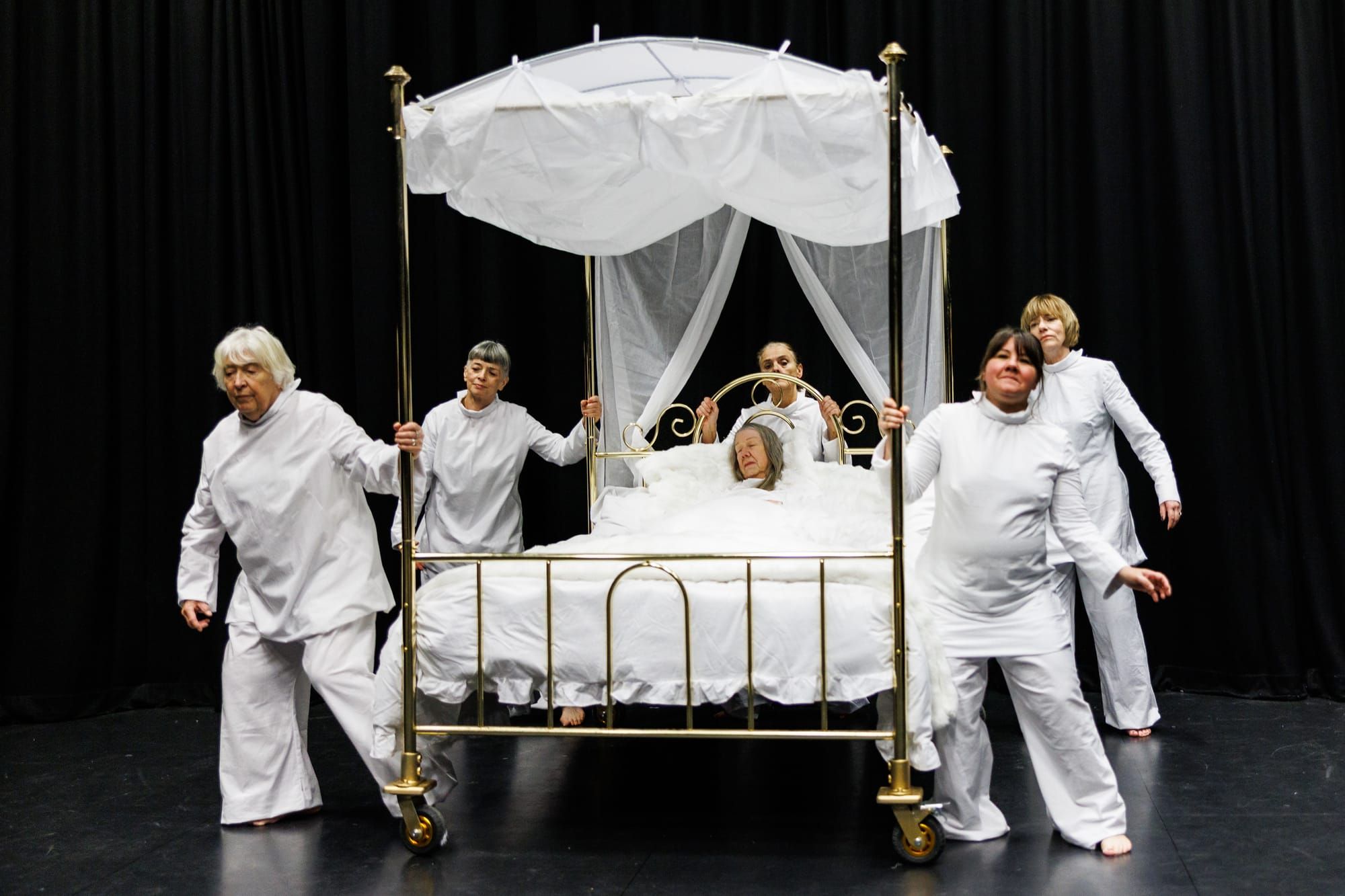 A group of women dressed in white stand around a gold four poster bed. Another woman is in the bed. 