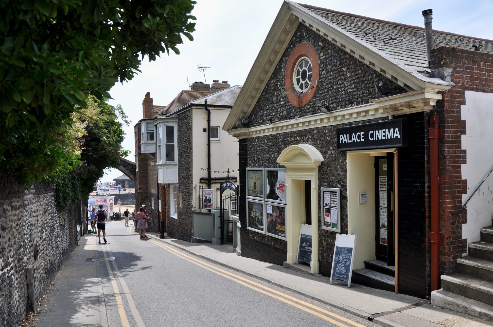 A view down a street to the sea. In the foreground is a small cinema housed in an old building. People mill around further down the street.
