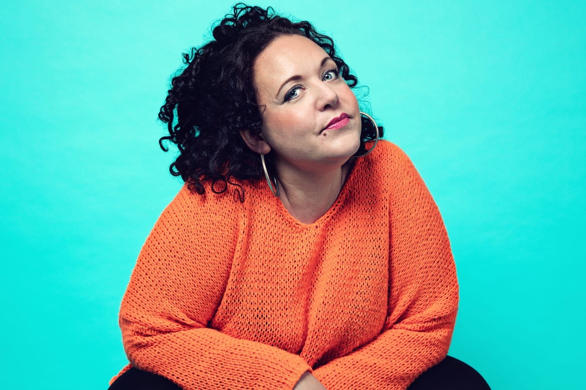 A woman with black hair, wearing hoop earrings, an orange knitted jumper and black trousers crouches with her head cocked to the side. 
