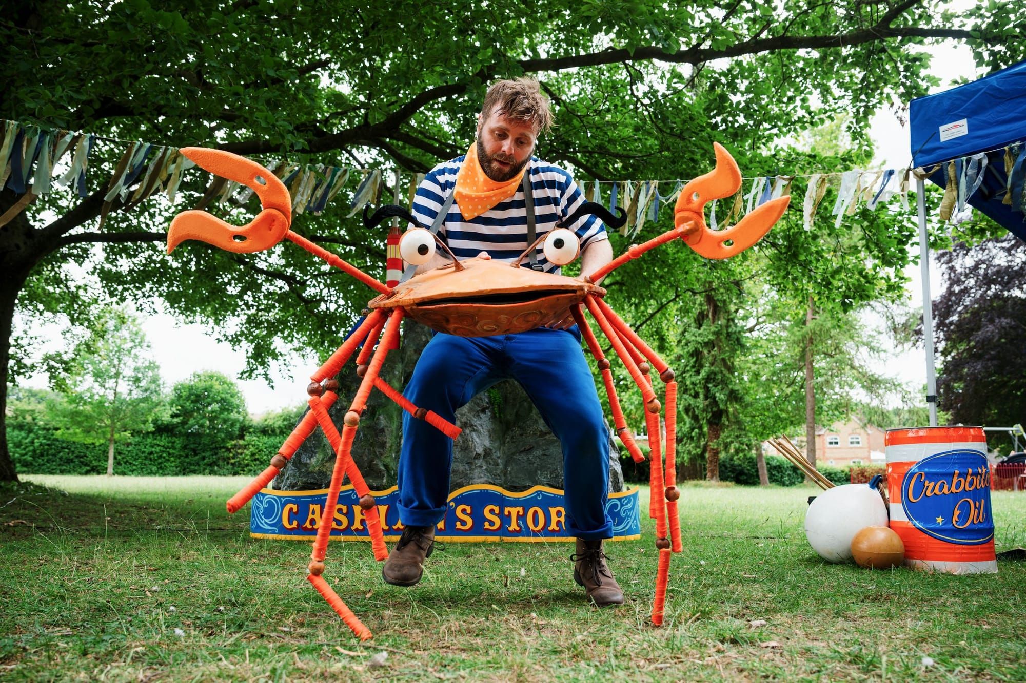 A man with brown hair dressed in blue and white operates an orange crab puppet standing on grass and in front of trees.