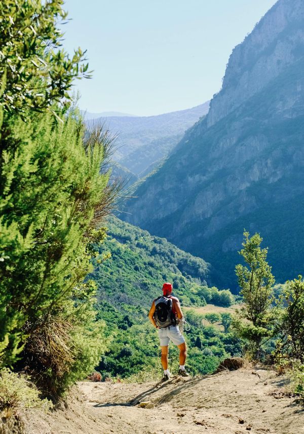 Man in orange shirt and white shorts standing on a rocky mountain trail with green hills and blue sky