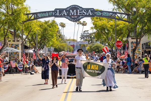 Historically dressed couple holding History Center banner marching down La Mesa Blvd. New La Mesa sign in the background.
