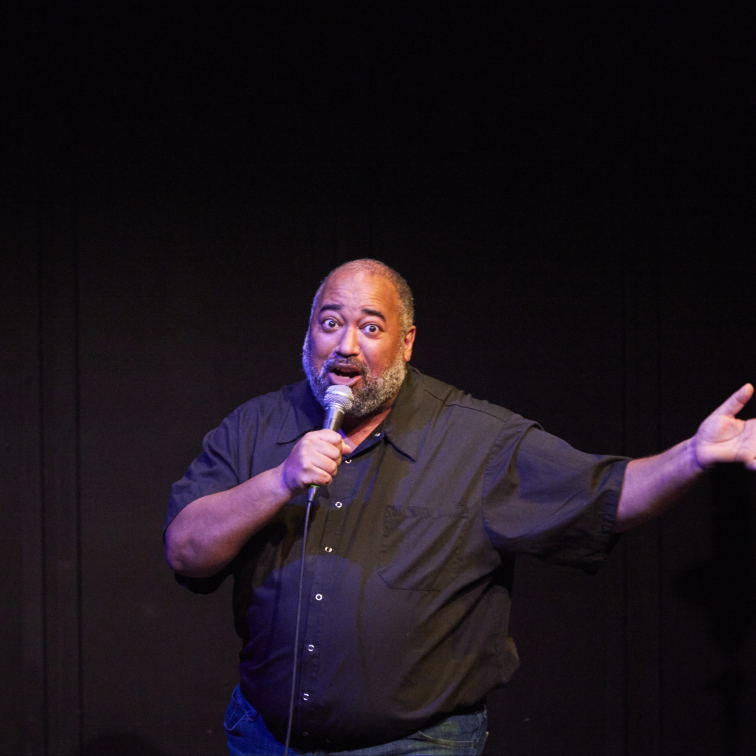 Velvet, a fat Black person of a certain age with salt & pepper hair, expansively extends their left hand out to the side while singing to the audience through a microphone.