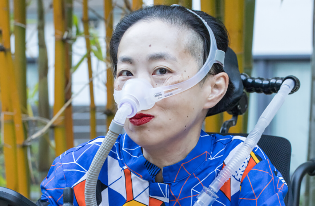 Photo of Alice Wong sitting in her wheelchair, with a mask over her nose to help her breathe. She's wearing red lipstick.