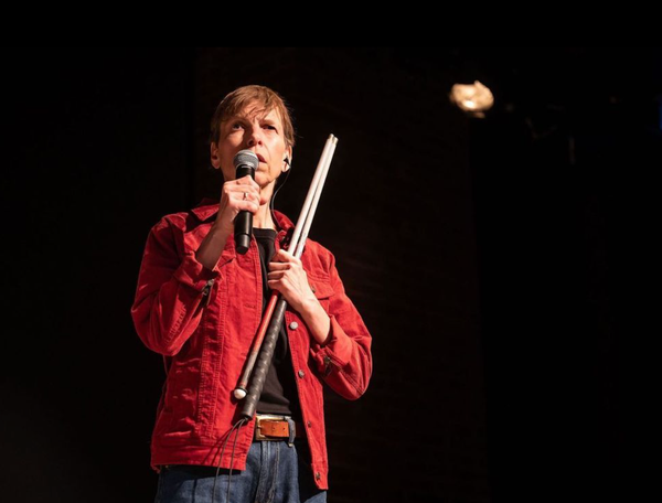 Alex Bulmer stands on stage with a microphone in one hand and her white cane clutched against her chest with her left arm.