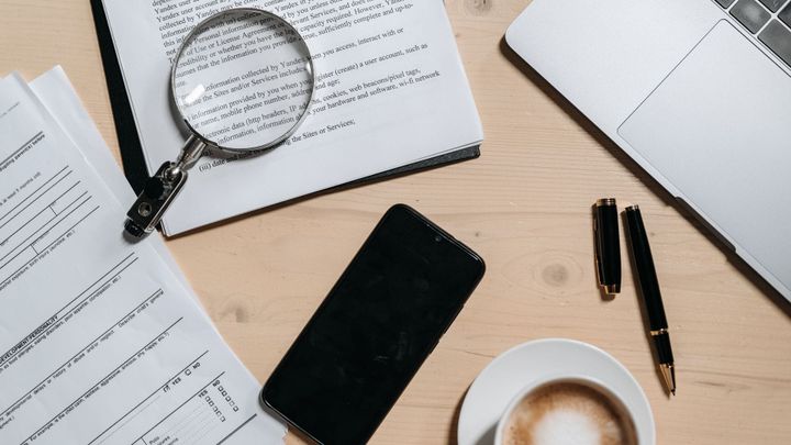Documents, a laptop, a magnifying glass, and a coffee cup on a desk.