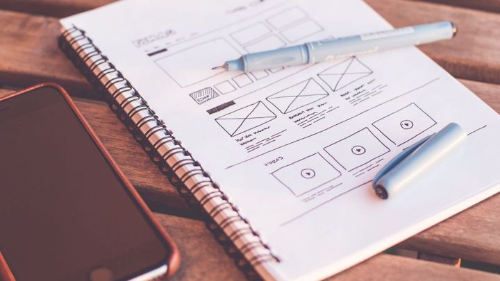 A notebook with wireframe sketches, a pen, and a smartphone on a wooden desk.