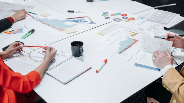 Paperwork, charts, markers, and office supplies spread across a white desk.