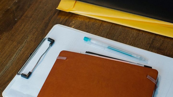 A notebook, clipboard, pen, and folders arranged on a wooden desk.
