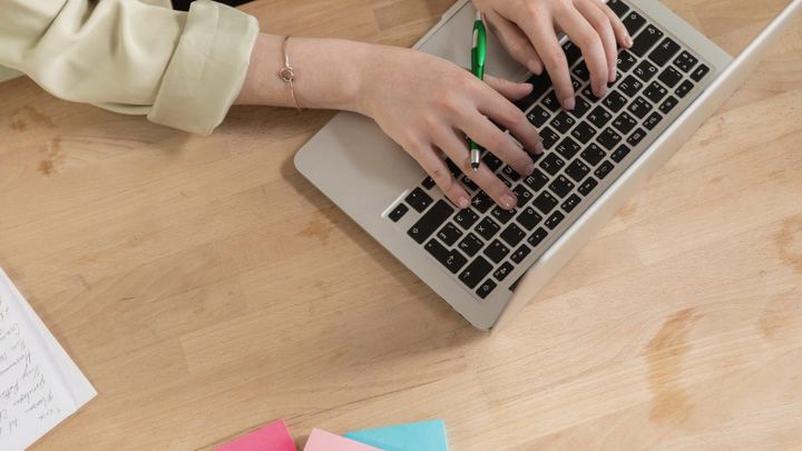 Hands type on a laptop on a wooden desk with a green pen and sticky notes nearby.