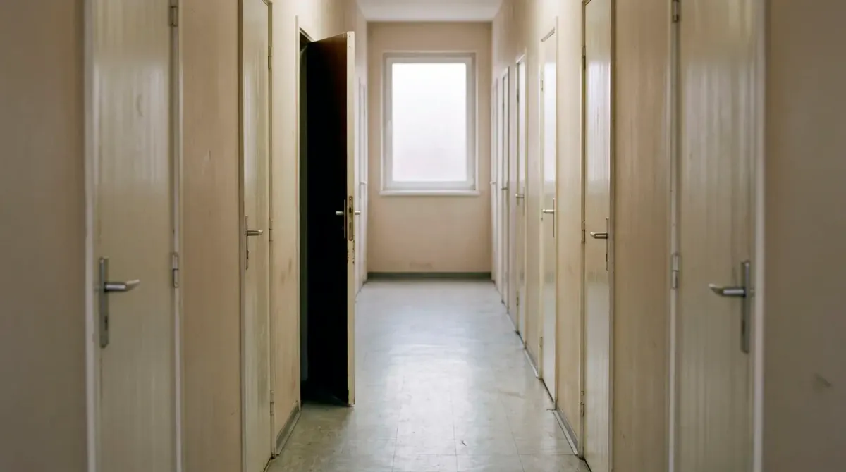 A plain corridor of identical closed doors in flat morning light, one of them slightly ajar with nothing visible beyond it, no people and no signage.