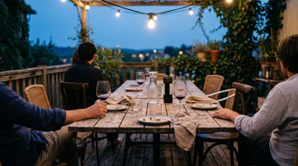A late-summer outdoor terrace at dusk with two small tables pushed together, half-empty glasses, a single overhead light, no faces visible.
