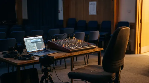 An empty operator's chair at a small mixing desk in a community hall, rows of empty chairs out of focus.