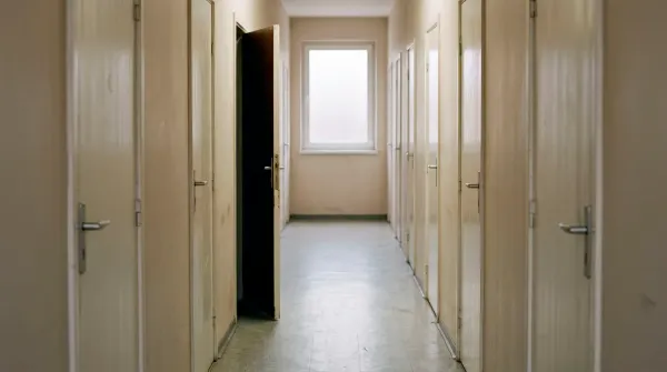 A plain corridor of identical closed doors in flat morning light, one of them slightly ajar with nothing visible beyond it, no people and no signage.