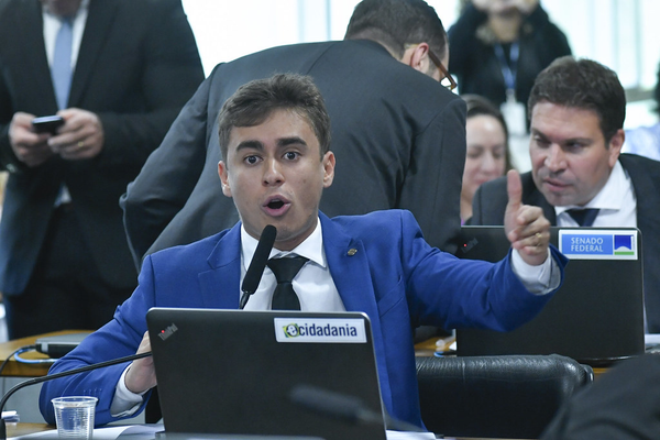 congressman nikolas ferriera at the brazilian senate in 2023. he's wearing a blue suit, black tie. he is pointing towards the camera with his fingers like a gun
