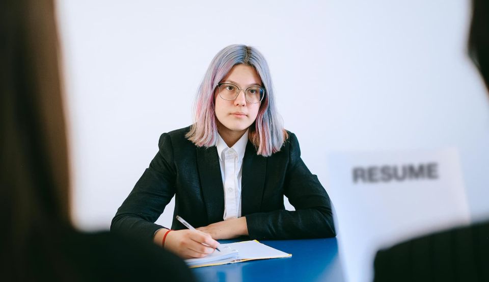 two interviewers, obscured, interview a woman with multicolor and mostly blue or pink or silver hair while holding a paper that says "resume"